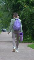 Rear view of an athletic woman in sportswear walking along park walkway after practicing outdoor yoga. Sportswoman in sportswear with backpack and exercise mat. Handheld shot, real time vertical video