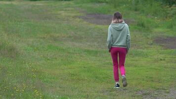 Full length view of unrecognizable athletic woman in sports clothing walking across field in short grass during summer sports training. Rear view of fitness model wearing hoodie and joggers. Zoom in video