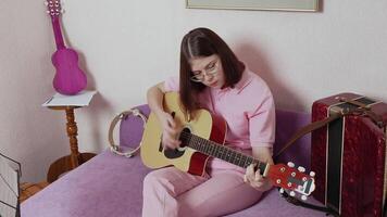 Young woman performs rendition on acoustic guitar in domestic studio setting. She is beginner guitarist and her practice is focused on refining technique and developing unique style. Handheld, zoom in video