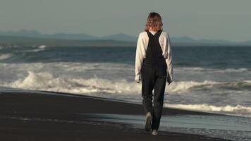Back view of unrecognizable fashion model walking on black sandy beach against backdrop of ocean breaking waves during summer beach holiday. Woman dressed in casual clothing. Full length, handheld video