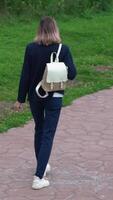 Rear view of an unrecognizable woman walking on footpath in public park in summer. Model wears blue jacket, jeans and carries small backpack on back. Handheld tracking shot, zoom in. Vertical video