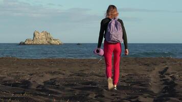 Rear view of unrecognizable female athlete with backpack and yoga mat walking on black sandy beach during sports training with ocean waves and island in background. Handheld shot, real time, zoom in video