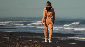 Unrecognizable woman walking black sandy beach on background of ocean breaking waves during summer vacation at golden hour. Full length rear view of model in orange jumpsuit. Real time , zoom in video