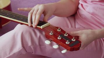 Closeup view of woman's hands tuning six string acoustic guitar. Hand moves methodically, tightening and loosening strings before playing. Guitarist sitting on sofa in home. Handheld, zoom in video
