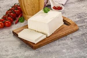 A piece of cheese on a cutting board next to tomatoes and a wooden bowl photo
