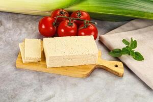 A piece of tofu on a cutting board next to a tomato and a piece of corn photo