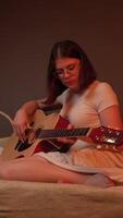 Young woman plays guitar with concentration on comfortable couch in living room. Scene is bathed in soft warm flickering light reminiscent of fireplace, creating an engaging atmosphere. Zoom in video