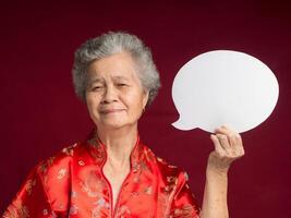 Senior woman wearing a traditional cheongsam qipao holding a blank speech bubble and with a smile while standing on a red background photo