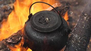 A well-used black kettle sits over an open fire, heating water in a rustic outdoor setting. Flames surround the kettle, creating a cozy campfire scene. video