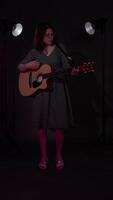 Musician strumming guitar, then curtseying, bending knees and bowing head at end performance to show gratitude from audience. Guitarist illuminated flickering multicolored lights at nightclub. Zoom in video