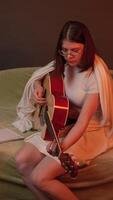 Young woman playing six string acoustic guitar with purposeful concentration, sitting on couch in living room highlighted soft and warm flickering light as if from a fireplace. Handheld shot, zoom in video