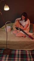 Young female guitarist is sitting on couch with guitar and reading musical notes. She is studying to practice playing guitar is illuminated by soft, warm flickering light like from fireplace. Zoom in video