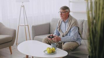 An elderly man in his 60s relaxes on a sofa at home, holding a game controller. He has grey hair and wears a light blue shirt with a sweater draped over his shoulders, enjoying his retirement. video