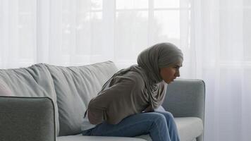A Middle Eastern woman wearing a hijab sits on a couch in her living room, visibly uncomfortable and holding her lower back. Sunlight filters through sheer curtains, creating a serene atmosphere. video