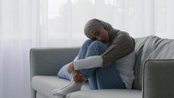 A woman dressed in a gray hijab sits curled up on a sofa in a sunny room, gazing thoughtfully out of a window. The soft lighting illuminates her peaceful expression. video