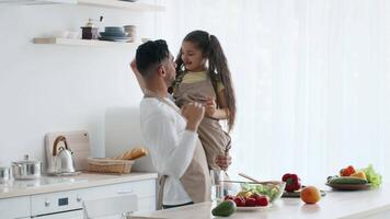 A joyful father lifts his daughter in the kitchen as they prepare a meal. The space is filled with fresh vegetables and fruits, creating a warm and inviting atmosphere. video
