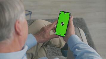 An elderly man in his 60s focuses on his smartphone while seated at home. He engages in a gaming activity, showcasing a relaxed retirement lifestyle during leisure time. video