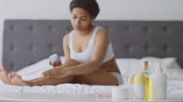 A young African American woman sits comfortably on her bed, applying skin care products as part of her relaxing beauty routine at home. She enjoys the self-care process in a calm environment. video