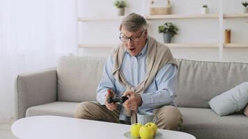 A senior man in his 60s relaxes at home, playing games while seated on a comfortable sofa. He has grey hair and is dressed casually, embodying a cheerful retirement lifestyle. video