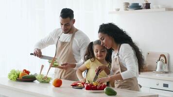 A family of three prepares a meal in a bright kitchen. The parents and their young daughter chop vegetables and share smiles while cooking together, emphasizing joy in family bonding. video