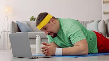 A man is engaged in a home workout, performing a plank on an exercise mat. He is focused on his laptop, following a fitness program, aiming for weight loss and better health. video
