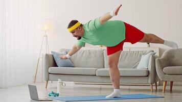 A millennial man with a chubby build engages in a home workout routine focusing on fitness and weight loss. He balances on one leg in a living room setting, following a virtual exercise session. video