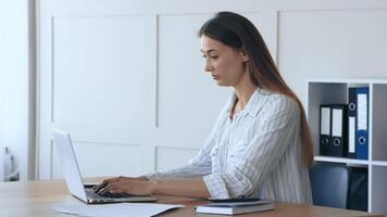 A woman is focused on her work at a laptop in a well-lit office. The setting features light-colored walls and organized shelves, creating a serene workspace conducive to productivity. video