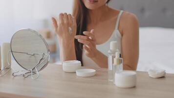 A young Arab woman applies moisturizer to her hand while seated at a wooden table. Various skincare products are neatly arranged before her, providing a relaxing and pampering atmosphere. video