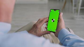 A senior man in his 60s sits comfortably in his home, engaged with a smartphone. He appears relaxed, enjoying his retirement lifestyle during the afternoon. video