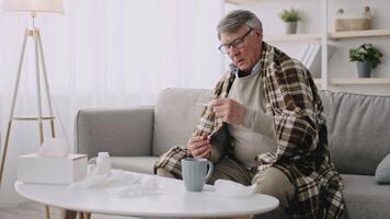 An elderly man in his 60s sits on a couch, wrapped in a blanket, looking discomforted. Soft light filters through the curtains, creating a calm atmosphere as he holds a cup and tissues nearby. video