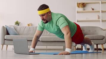 A man focused on his workout routine is seen doing push-ups at home. He wears a green shirt, red shorts, and headband, following a fitness routine displayed on his laptop. video