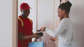A man with a red cap and backpack is delivering packages to a woman at her apartment door. They are smiling and engaged in a friendly conversation, showcasing a modern delivery service encounter. video