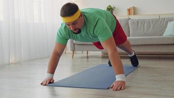 A man in a green shirt and red shorts works out on a blue mat in his home. He focuses on a fitness routine to lose weight, displaying determination and effort in a sunny indoor setting. video