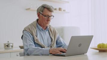 An elderly man in his 60s uses a laptop while sitting at a kitchen table. He appears focused on his online task, surrounded by a light and airy home environment. video