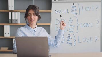 A young woman teaches an online class from her home, smiling as she interacts with students. A whiteboard with lesson notes is visible in the background, highlighting her teaching style. video