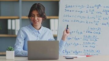 A young teacher is leading a remote math lesson using a laptop at home. She engages with students while explaining concepts on a whiteboard filled with equations. video