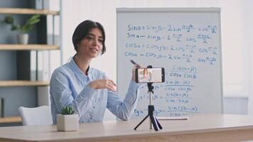 A young woman conducts an online lesson, using her phone to interact with students. Mathematics concepts are illustrated on the whiteboard behind her while she engages the audience with enthusiasm. video