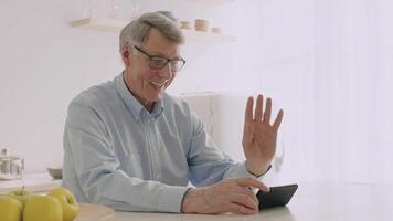 An elderly grey-haired man in his 60s smiles and waves during a call at home. He sits comfortably at a kitchen table, surrounded by apples and a bright, cheerful environment. video