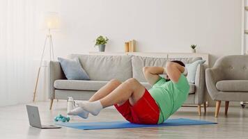 A man in workout clothes is lying on a mat, doing exercises for fitness and weight loss. He focuses on his routine, with a laptop beside him for guidance. video