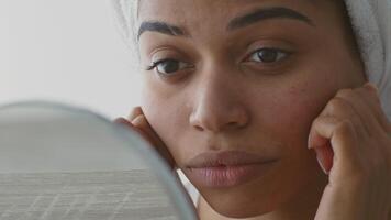 A young African American woman engages in her skincare routine at home, looking closely in the mirror while taking time for self-care and relaxation. She appears focused and serene. video