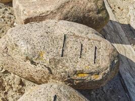 A closeup view of a large, weathered rock with distinct markings on its surface. The rock is surrounded by other stones and has some yellow lichen growth. The markings appear to be carved lines photo
