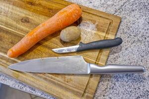 A wooden cutting board with a large chef's knife, a small paring knife, a carrot, and a potato. The scene is set in a kitchen environment, showcasing fresh vegetables and kitchen tools. photo