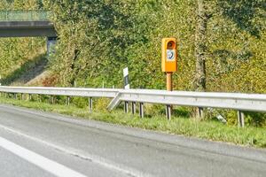 A roadside view featuring a speed camera mounted on an orange pole, with a highway and green trees in the background. The road is clear and well-maintained, indicating a rural or suburban area. photo