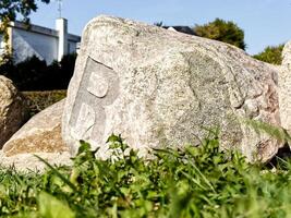 A large stone with the letter R carved into it, surrounded by grass. The stone is weathered and textured, set against a backdrop of greenery and a building in the distance. photo