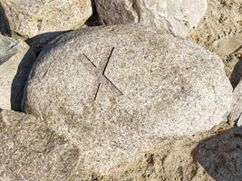 A large, smooth rock with a carved X symbol on its surface, surrounded by smaller rocks and sand. photo