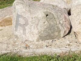 A large, weathered rock with the letter R engraved on its surface, surrounded by smaller stones and sandy ground, with green grass in the foreground. photo