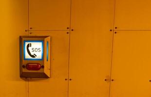 An emergency SOS call box mounted on a wall, illuminated with blue light. The box features a phone icon and the letters 'SOS' prominently displayed. photo