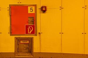 A bright yellow wall featuring a fire hydrant access door and a fire alarm. The door is marked with the number 5 and includes a fire hydrant symbol. The overall color scheme is warm and industrial. photo