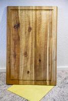 A wooden cutting board with a smooth surface and natural grain patterns, placed on a light-colored carpet next to a yellow cloth. The board features a rectangular shape with a decorative edge. photo