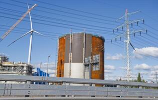 A large industrial structure with a cylindrical shape, surrounded by wind turbines and power lines. The scene captures a mix of renewable energy and industrial development under a clear blue sky. photo